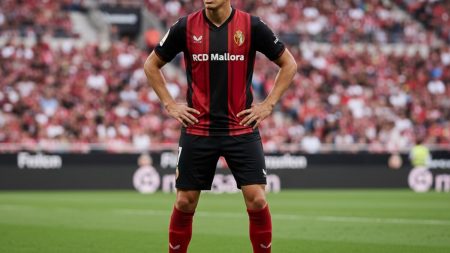 Fußballspieler in rotem RCD Mallorca-Trikot im modernen Stadion, steht konzentriert im Mittelpunkt und symbolisiert Spielserie und Konstanz