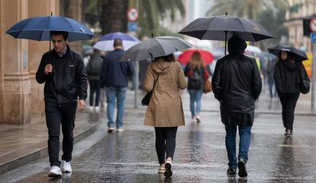 Regnerisches Wetter in Palma de Mallorca mit Personen unter Regenmänteln und Schirmen auf der Straße.